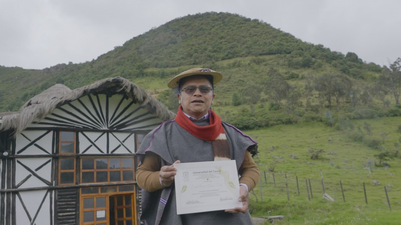 A male Agroecology course graduate smiles and holds up his certificate, whilst stood in front of a black and white building and lush green hillside.